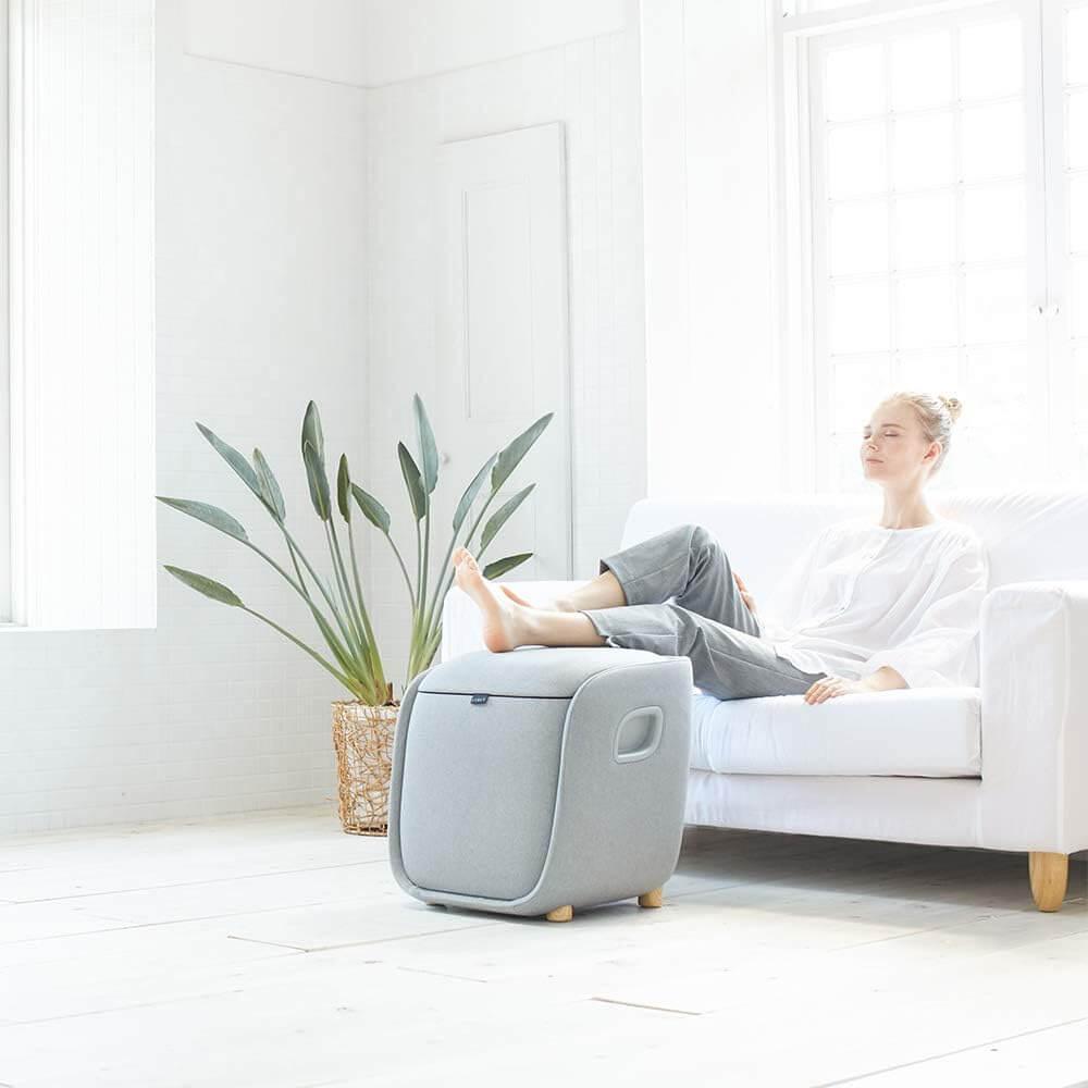 Synca Wellness REI Foot Massager in gray with a woman relaxing on a white couch using the electric massager for foot therapy