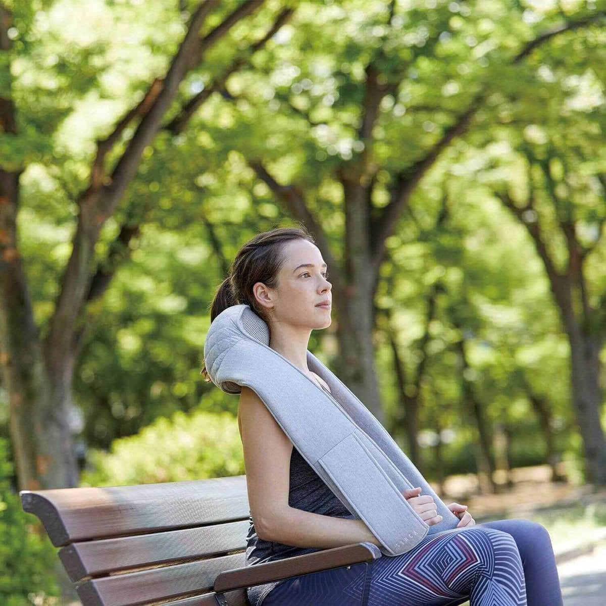 Neck and Shoulder Massager in light gray fabric, worn around a seated woman's neck with padded shoulder arms.